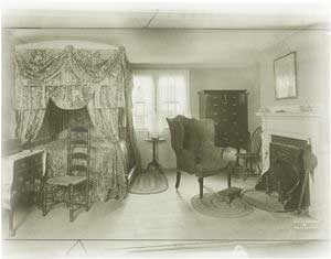 A New England bedroom of about 1800, showing four poster with curtains and valance, and truckle bed to be pulled out from beneath larger bed.